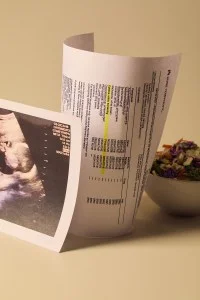 Three items are arranged on a white surface: a copy of a sonogram, a printout of positive drug test results, and a salad in a white bowl.