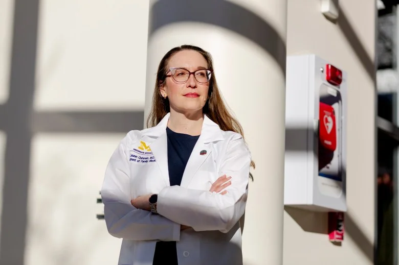 A White female doctor poses for a portrait at a hospital. She is wearing glasses, a black blouse, and a white lab coat.