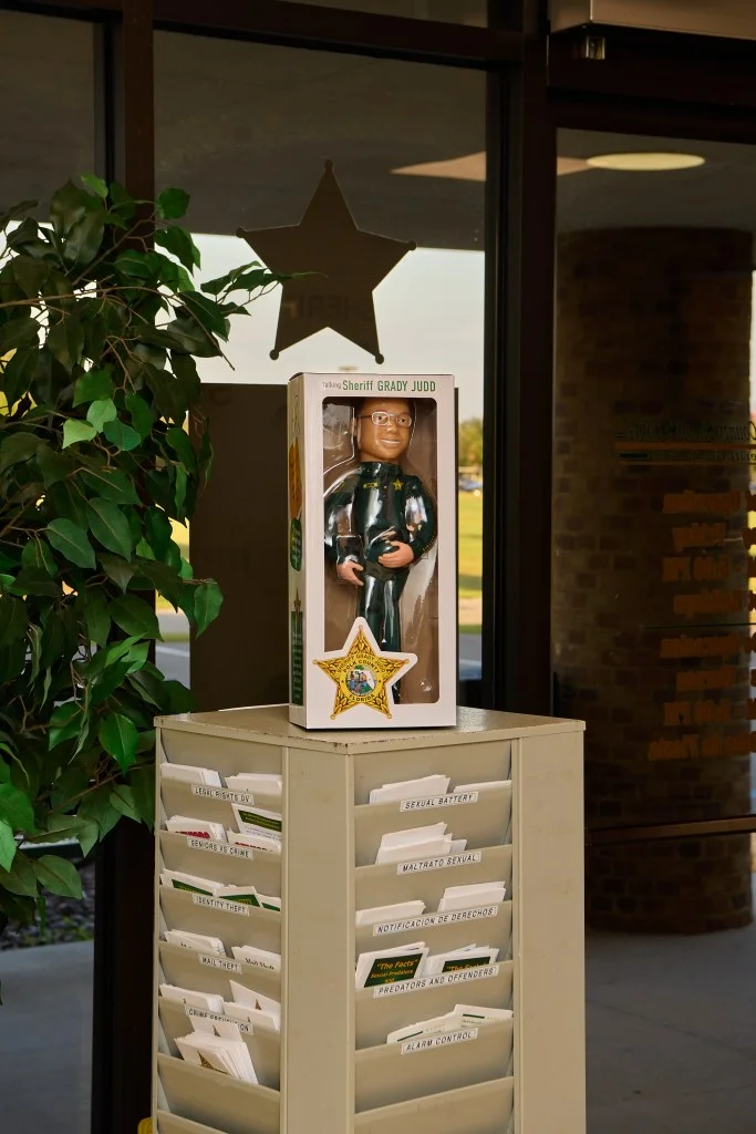A doll with glasses and in a sheriff's uniform is in a box, sitting on top of a filing cabinet. You can see the star of the sheriff's office badge in the window behind it.