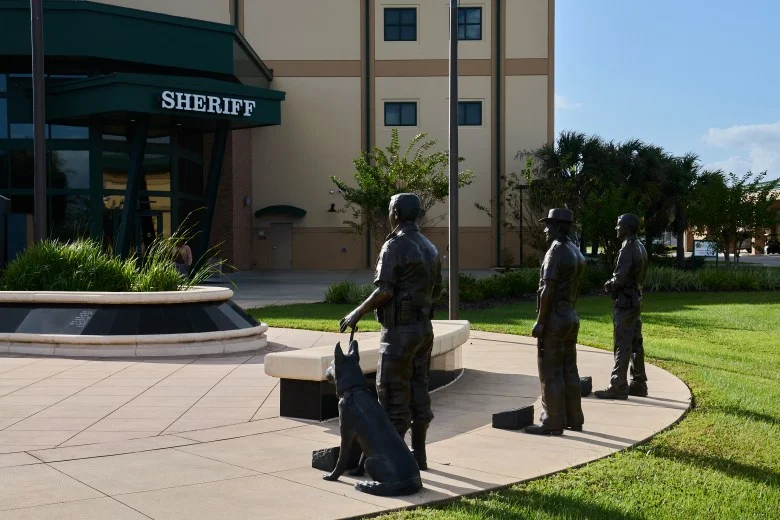 Three bronze statutes representing law enforcement officers, one with a K-9 officer, stand in a ring along green grass and outside a building marked Sheriff on the awning.