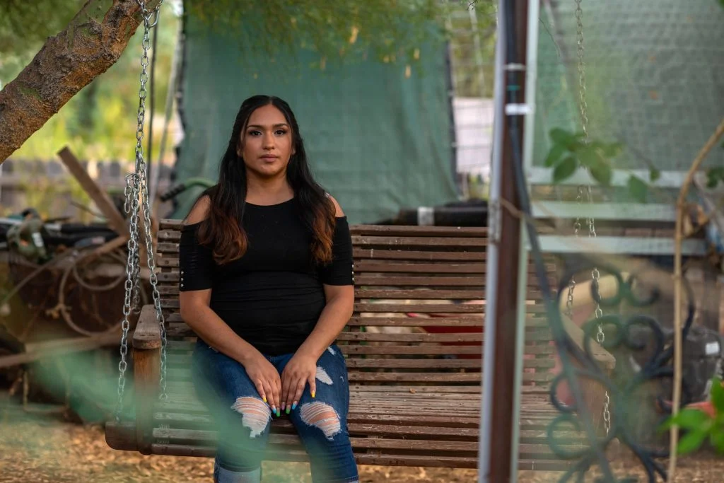 A woman with long dark hair sits on a bench swing. She wears a black short-sleeve top with shoulder cutouts and dark ripped jeans.