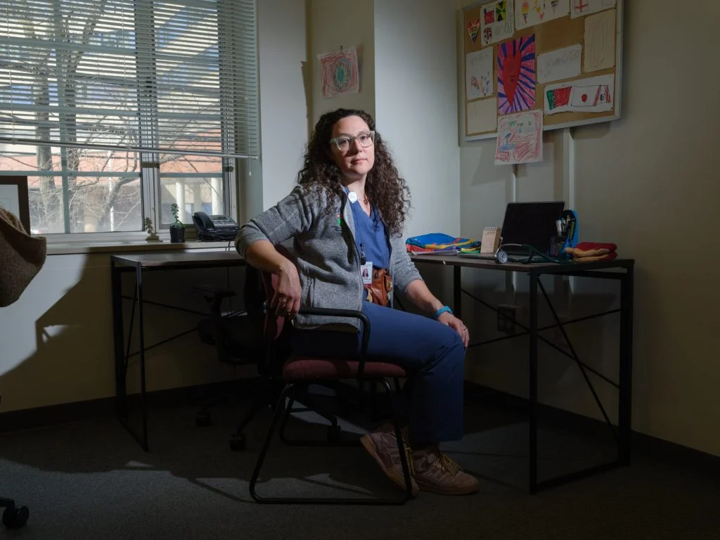 A White female doctor, wearing glasses, a gray jacket, and blue scrubs, sits at her desk. Children's hand drawings hang on the wall.