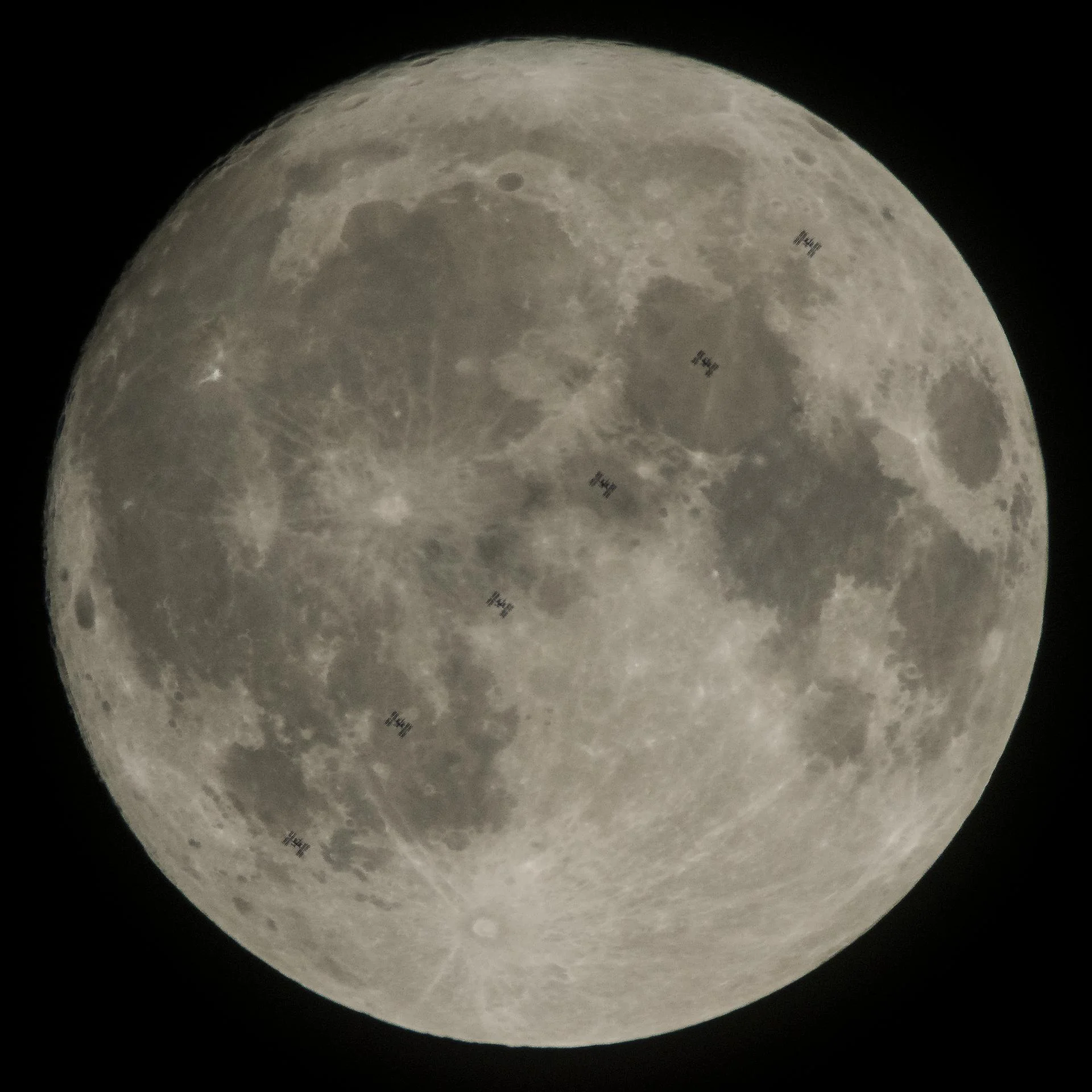 The full Moon fills the frame against a black sky, with multiple small silhouettes of the International Space Station captured as it transits across the Moon’s face.