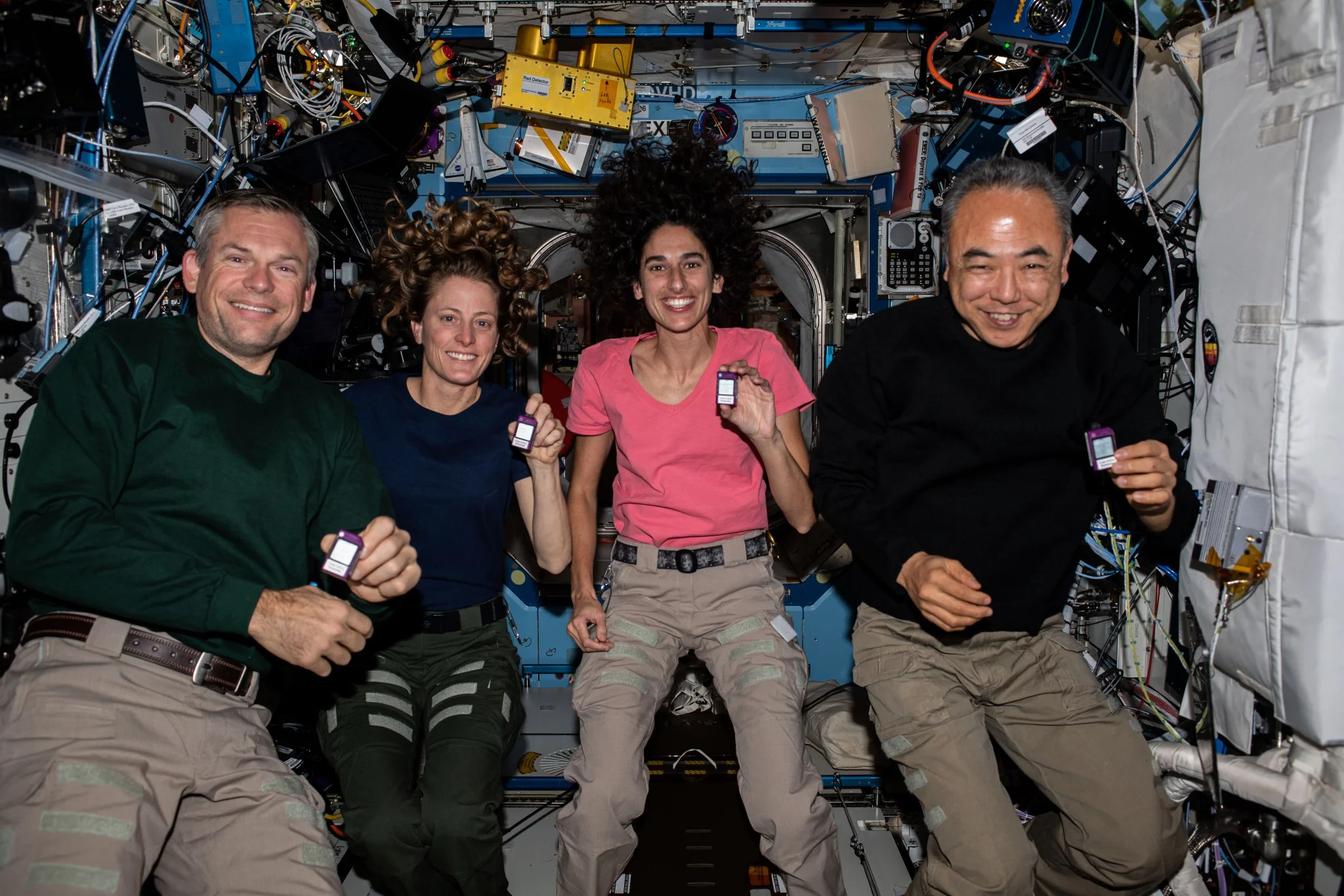 Four astronauts float together inside the International Space Station, smiling and holding small devices about the size of a printer ink cartridge. They are surrounded by equipment, cables, and modules in the microgravity laboratory.
