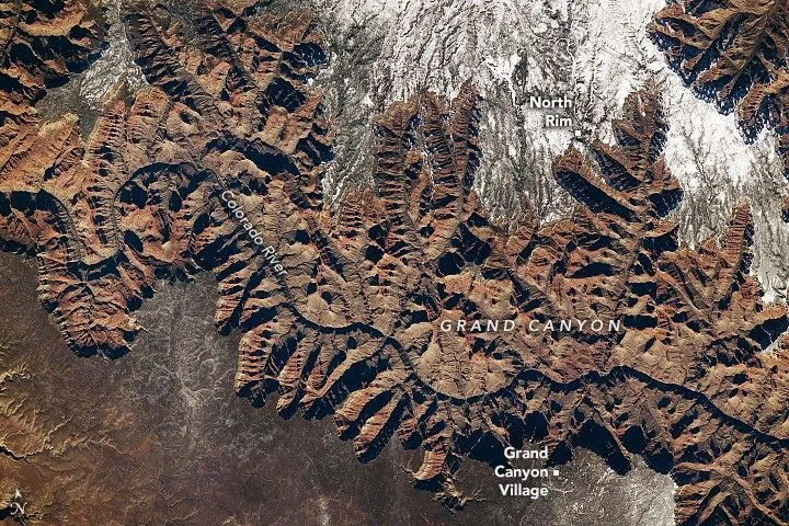 A portion of the Grand Canyon, with the curving Colorado River at its bottom, runs across the width of this downward-looking photo. Snow covers some of the plateau on both sides of the canyon, with more appearing on the North Rim.